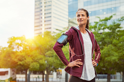 Latin sport woman listening music with earphones while standing and resting after running outdoors. Mature woman wearing jacket and standing after morning run in urban city. Satisfied runner looking away with pride.
