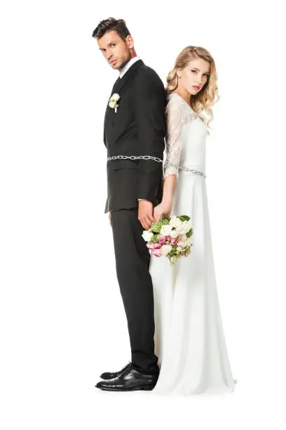 serious young newlyweds tied with chain back to back and looking at camera isolated on white