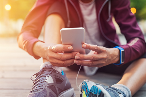 Closeup sporty woman hand using smartphone while sitting on floor. Woman wearing jacket and typing on mobile phone to check pulse rate in the early morning. Runner hand using smart phone while sitting on ground after running.