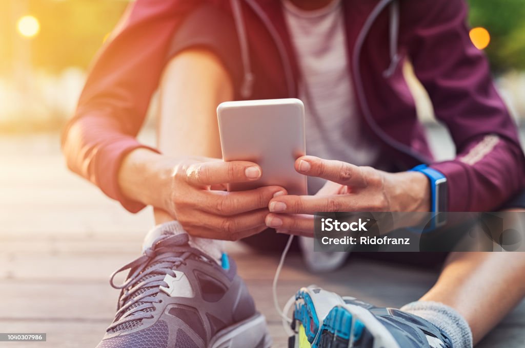 Sporty woman using mobile phone Closeup sporty woman hand using smartphone while sitting on floor. Woman wearing jacket and typing on mobile phone to check pulse rate in the early morning. Runner hand using smart phone while sitting on ground after running. Exercising Stock Photo