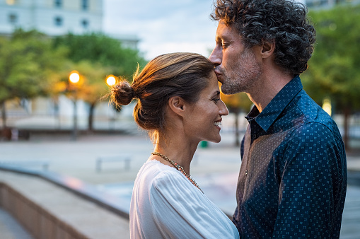 Mature husband kissing wife on forehead in the street in the evening. Romantic senior man giving a kiss to her woman in the city street. Loving middle aged couple in love at dusk.