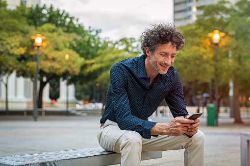Mature businessman sitting on bench and using smart phone at dusk. Happy smiling guy in smart casual reading a message on cellphone. Stylish man surfing the net with smart phone in the evening.