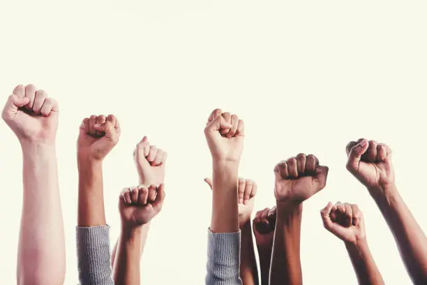 A multi-ethnic group of hands are raised giving Black power clenched-fist salutes on a white background with ample copy space.