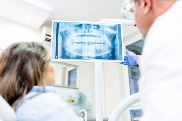 Photo of Dentist showing patient x-ray Photo of health dental professional in blue scrubs examining dental x-ray on computer screen, isolated dentist office. People, medicine, technology and health care concept - dentists looking to x-ray scan on monitor at dental clinic. Rear view of male dentist explaining the details of x-ray to a beautiful female patient digital xray stock pictures, royalty-free photos & images