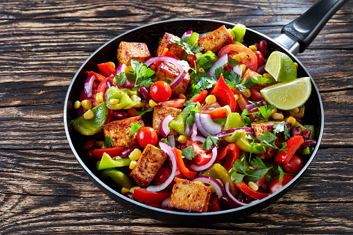 Delicious Cowboy Caviar cooked from colorful, fresh vegetables: green and red pepper, black beans, sweet corn, tomato, red onion, tofu cheese, garnished with parsley leaves, on a black skillet