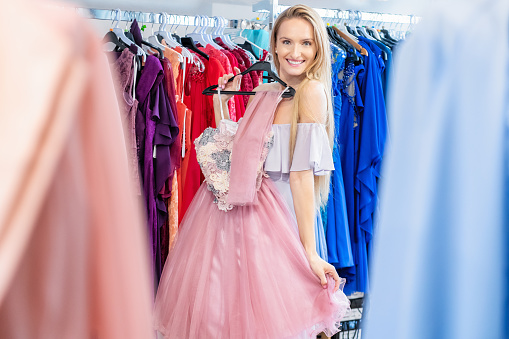 Beautiful woman shopping and choosing the right dress for luxury event. Young woman holding and trying on elegant gown and smiling at camera. Focus on background.