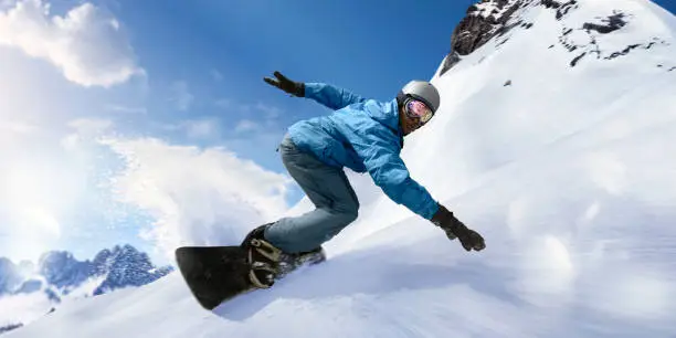 A close up image of a black snowboarder laying into a carve turning the snowboard towards the camera. The snowboarder is dressed in blue jacket, salopettes, helmet and visor and is travelling at high speed down a slope with snow spraying out from behind the board. With motion blur.
