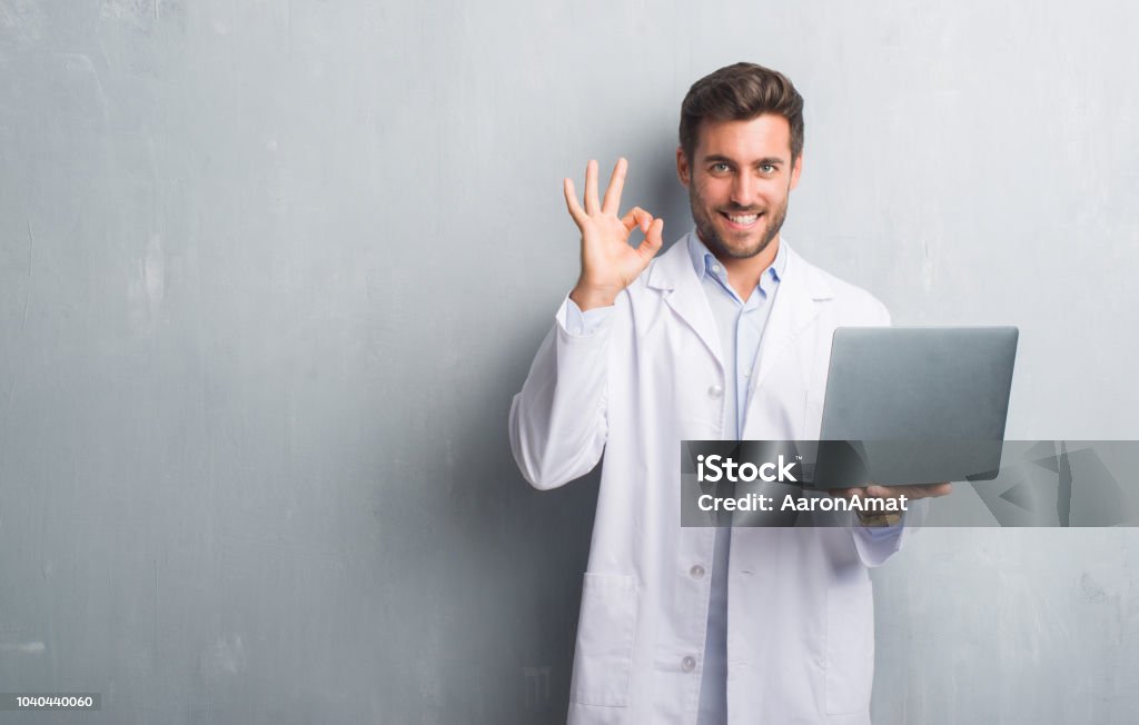 Handsome young man over grey grunge wall wearing white coat using laptop doing ok sign with fingers, excellent symbol Adult Stock Photo