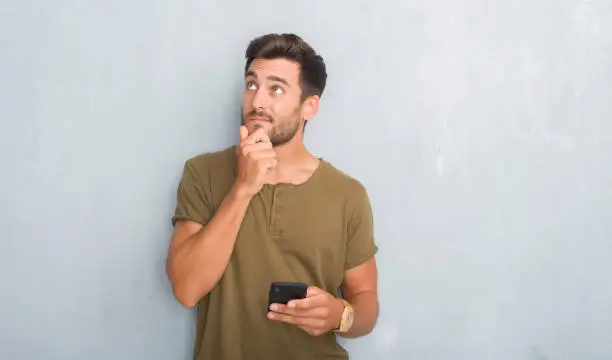 Photo of Handsome young man over grey grunge wall texting a message using smartphone serious face thinking about question, very confused idea