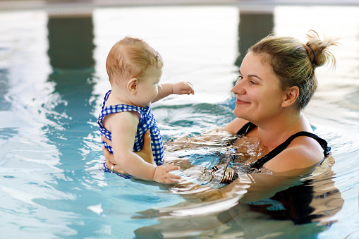Happy mother swimming with cute adorable baby daughter in swimming pool. Smiling woman and little child, girl of 6 months having fun together. Active family spending leisure and time in spa hotel.