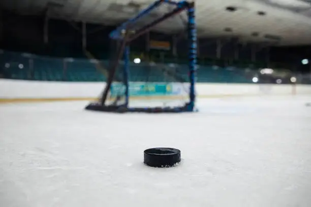Low angle view of a ice hockey puck stationary on the ice infront of a net on the ice rink. The ice rink has no people in and the seats are empty.