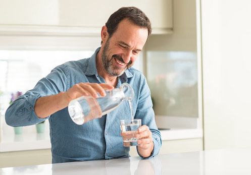 Hombre de mediana edad beber un vaso de agua con una cara feliz de pie y sonriendo con una sonrisa de confianza mostrando los dientes photo