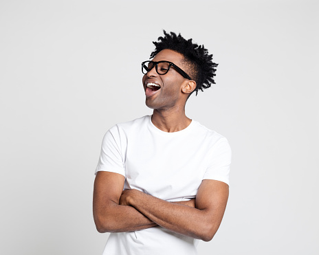Portrait of afro american man with surprised expression, looking away at copy space and laughing on white background. Man in white t-shirt wearing nerd glasses with arms crossed.