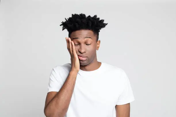 Close up studio portrait of afro american man with toothache against white background. Young man touching mouth with hand with painful expression.