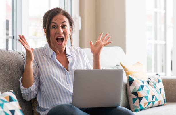 istock Middle aged woman using laptop in the sofa very happy and excited, winner expression celebrating victory screaming with big smile and raised hands 1040481002