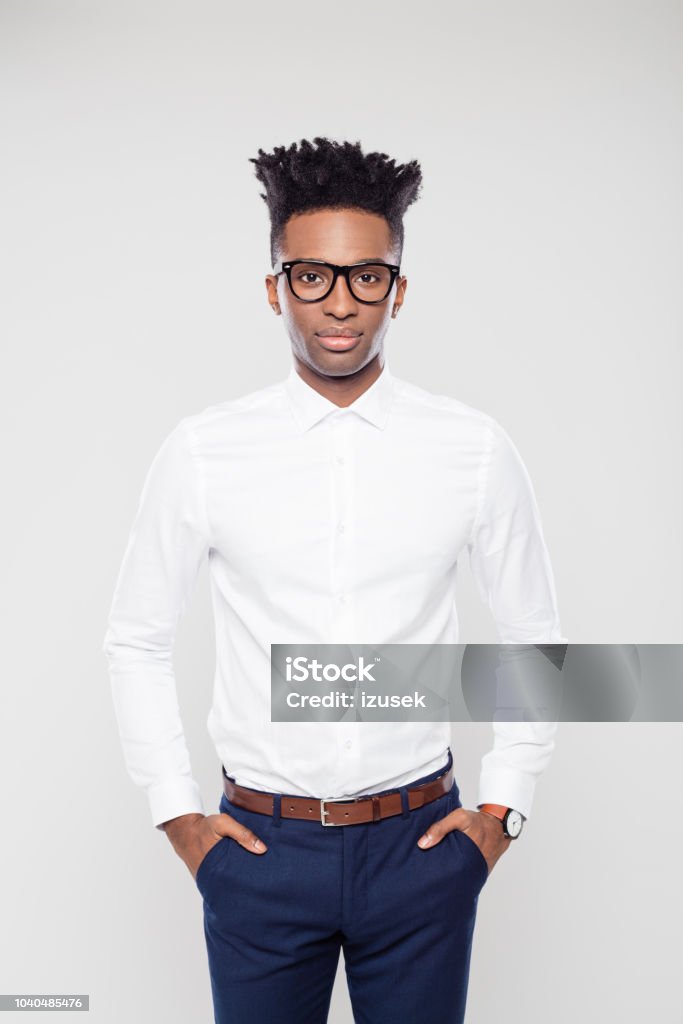Handsome businessman with his hands in pocket Portrait of handsome afro american businessman standing with his hands in pocket on white background. African guy in formal wear wearing eyeglasses staring at camera in studio. Button Down Shirt Stock Photo