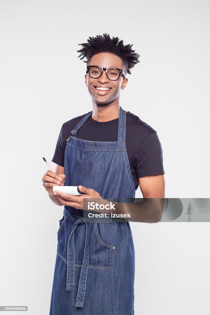 Happy waiter with pen and notepad Portrait of happy young waiter with pen and notepad looking at camera and smiling. Afro american man wearing blue apron taking order. Barista Stock Photo