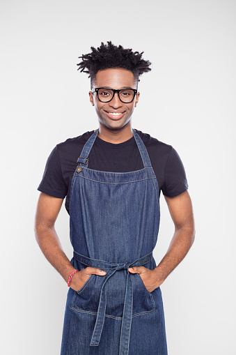 Portrait of young afro american male shop worker in apron standing with his hands in pocket on white background. Male barista wearing apron and eyeglasses looking at camera.
