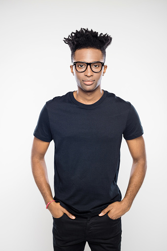 Portrait of handsome afro american man wearing black t-shirt and jeans standing with his hands in pocket staring at camera. Young man with funky hairstyle and nerd glasses on white background.
