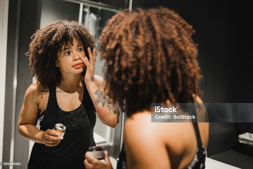 Taking Care of Her Skin Young woman applying skin moisturiser to her face. She is standing in the changing rooms in the gym and looking in the mirror. Skin Care Stock Photo