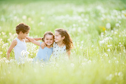 Three multi ethnic siblins hold onto each other as they laugh and walk through a meadow of tall grass and dandelion flowers.