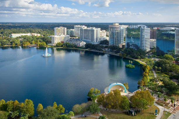 Lake Eola Surrounded by Buildings and Trees A fountain and two lush palm trees in the middle of Lake Eola. Cityscape surrounding the lake emphasizing the cohabitation of man and nature in Florida. A large walkway for tourists and locals to walk around the lake, and a background of trees, clouds, and sky encompass the rest of the shot. orlando florida stock pictures, royalty-free photos & images