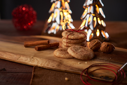 Cinnamon sticks and walnuts surround these snickerdoodles wrapped and unwrapped in a thin red bow on a wooden plank. Two lighted, metal, blurry Christmas trees with a red tinsel ball are in the background.