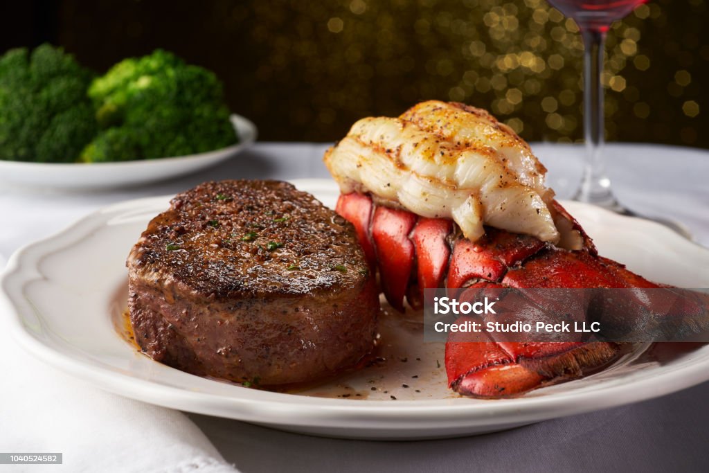 Steak and Lobster on a White Plate and Table Cloth A juicy steak next to a succulent lobster in a white fancy restaurant. There is a plate of blurry broccoli in the background, and this delicious meal is paired with a red wine in a wine glass shown to the right. There is light in the background bokeh into octagons. Steak Stock Photo