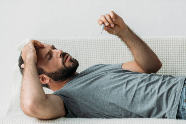 istock sick bearded man lying on couch and holding thermometer 1040532308
