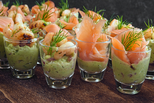 cold buffet with appetizers in glasses, prawn shrimps and salmon on avocado cream and dill garnish on dark wood, selected focus, narrow depth of field