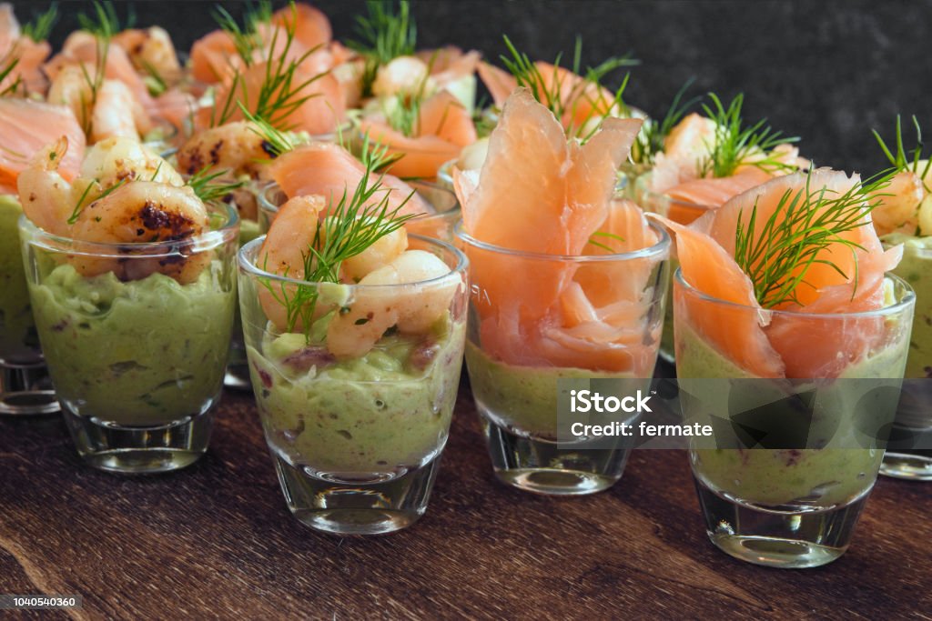 cold buffet with appetizers in glasses, prawn shrimps and salmon on avocado cream and dill garnish on dark wood cold buffet with appetizers in glasses, prawn shrimps and salmon on avocado cream and dill garnish on dark wood, selected focus, narrow depth of field Buffet Stock Photo