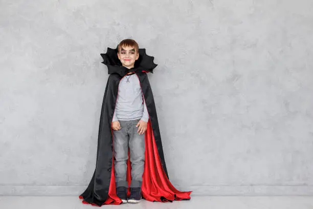 Smiling boy with vampire makeup and black, red cloak with bat like collar on a gray textured background. Halloween mood, copy space available. Cute kid wearing halloween costume.