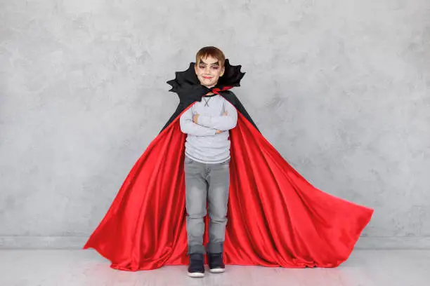 Smiling boy portrait wearing Halloween vampire makeup with fluttering red, black cloak, on a gray textured background. Cute kid wearing super hero costume with crossed hands, copy space available.