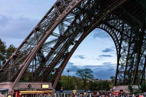 Paris, France - May 9, 2013: People standing in line on the pavement underneath the Eiffel Tower - waiting to visit the famous Parisian Landmark built in 1889 for the Exposition Universelle. The park Champ de Mars is seen in the background.