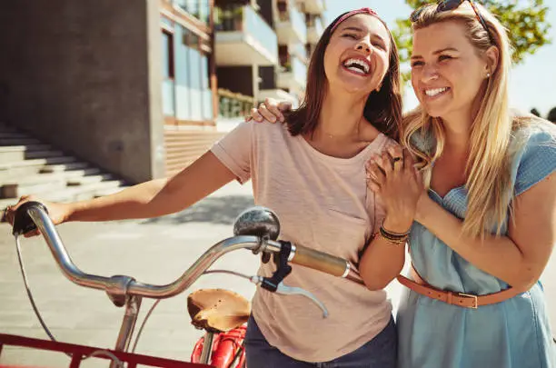 Two young female friends walking through the city with their bike laughing and having fun together