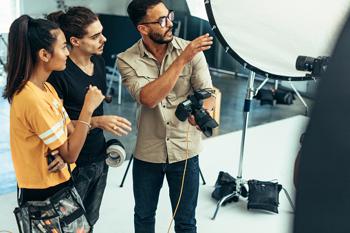 Photographer explaining about the shot to his team in the studio. Photographer talking to his assistants holding a camera during a photo shoot.