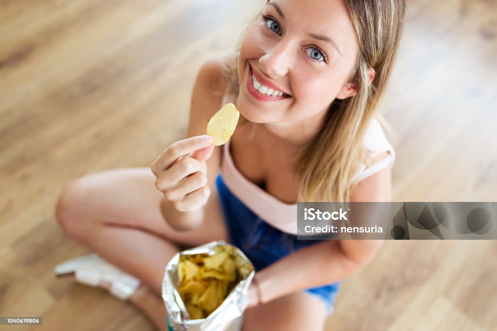 Beautiful young woman eating patatoes while sitting on the floor at home. Portrait of beautiful young woman eating patatoes while sitting on the floor at home. Potato Chip Stock Photo