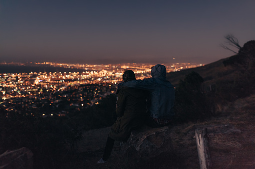 Couple watching the brightly lit city sitting on top of a hill late in the evening. Rear view of couple sitting together on a hill talking and enjoying the city view at night.
