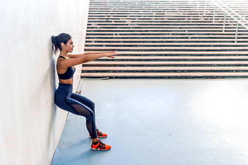 Photo of sporty athletic woman in sneakers and tracksuit squatting doing sit-ups while leaning on the wall during the day.