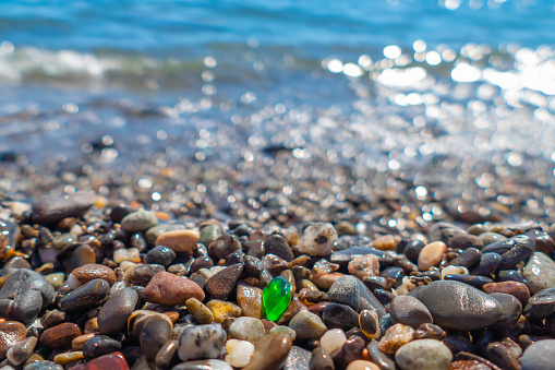 Small green polished glass stone among pebbles on sea beach on sunny summer day