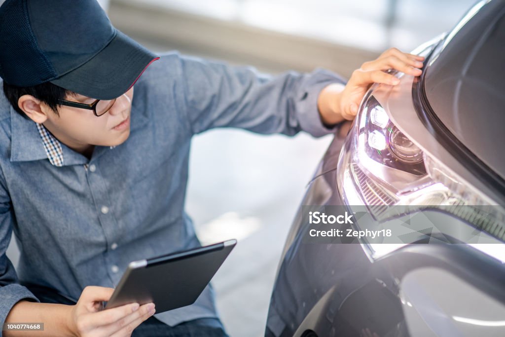 Young Asian auto mechanic holding digital tablet checking headlight in auto service garage. Mechanical maintenance engineer working in automotive industry. Automobile servicing and repair concept Headlight Stock Photo