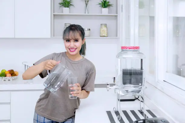 Beautiful young woman smiling at the camera while pouring fresh water into the glass in the kitchen room at home