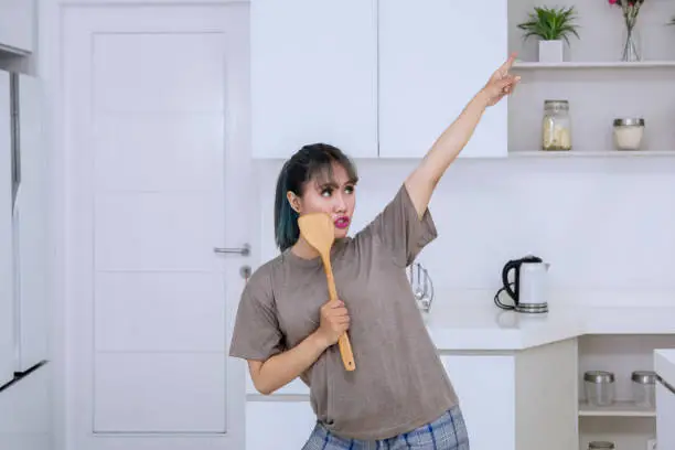 Beautiful young woman dancing and singing with a wooden spatula as a microphone in the kitchen room at home