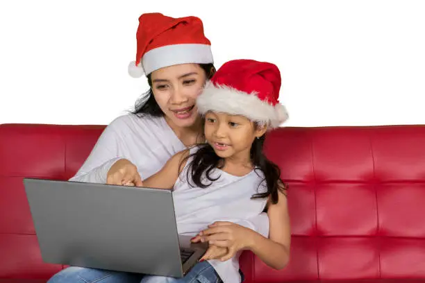 Picture of young mother and her daughter wearing Santa hat while using a laptop, isolated on white background