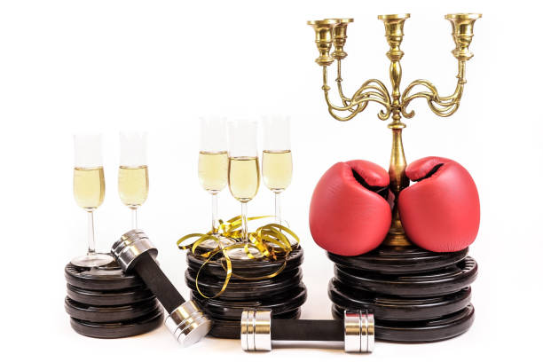 New Year Resolution. Celebrate new years eve at the gym. Glasses of champagne on weight plates and dumbbells. Boxing gloves on candelabra. New Year Resolution. Celebrate new years eve at the gym. Glasses of champagne on weight plates and dumbbells. Boxing gloves on candelabra. Workout and exercise more next year. White background. 2019 new year resolution stock pictures, royalty-free photos & images