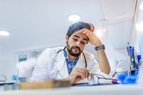 Stressed male doctor sat at his desk Overworked doctor in his office. Tired male scientist trying to focus, rubbing his forehead with fingers. Mid adult male doctor working long hours. Stressed male doctor sat at his desk head in hands photos stock pictures, royalty-free photos & images