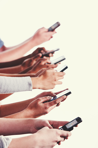 A multi-ethnic group of hands are raised against a white background, each holding a smart phone and busy sending a text.