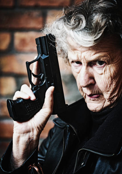 Fierce old woman holding gun raises eyebrow in warning Wrinkled old woman holds up a semi-automatic pistol and raises one eyebrow, staring at the camera confidently, warning of her ability to defend herself. old guns stock pictures, royalty-free photos & images