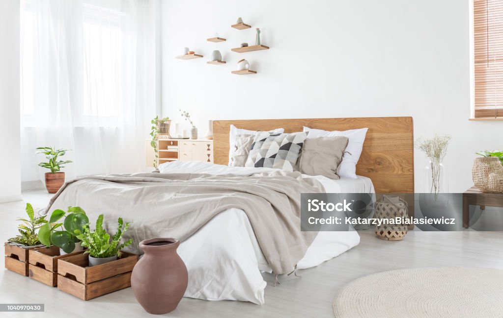Pillows and sheets on wooden bed in bright bedroom interior with plants and windows. Real photo Bedroom Stock Photo