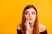 Close up portrait of  young girl tells a gossip secretly looking away and putting her hand to her mouth isolated on yellow background